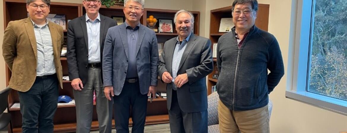  Five men are standing together in an office, smiling at the camera. They are dressed in business casual attire, and behind them is a bookshelf with various items, including plants and books.
